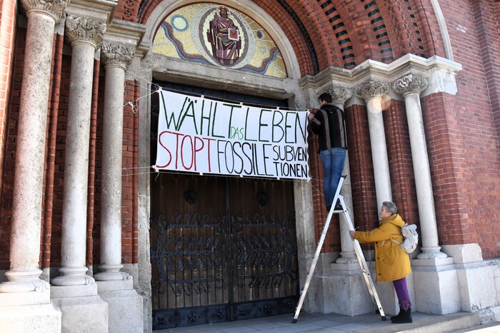 Ein Banner mit der Aufschrift “Wählt das leben - Stopt fossile Subventionen.” wird an einer Kirchentüre angebracht.