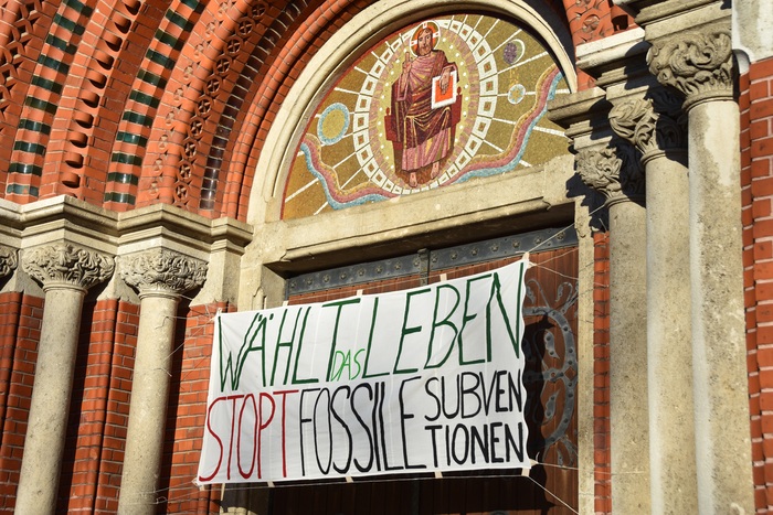 Ein Banner mit der Aufschrift “Wählt das leben - Stopt fossile Subventionen.” hängt an einer Kirchentüre.
